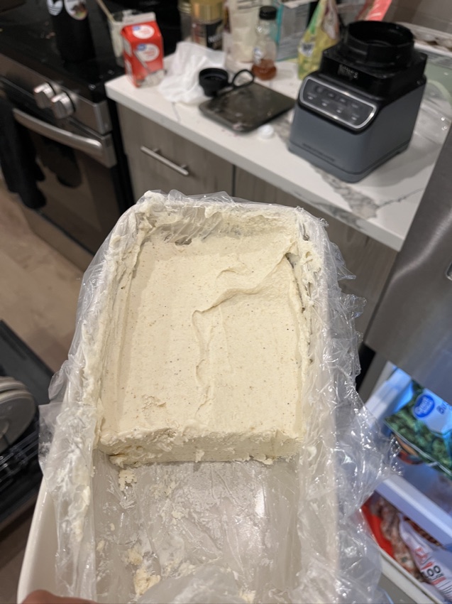 A tub of homemade malai kulfi in a plastic-lined tin, held up in a kitchen